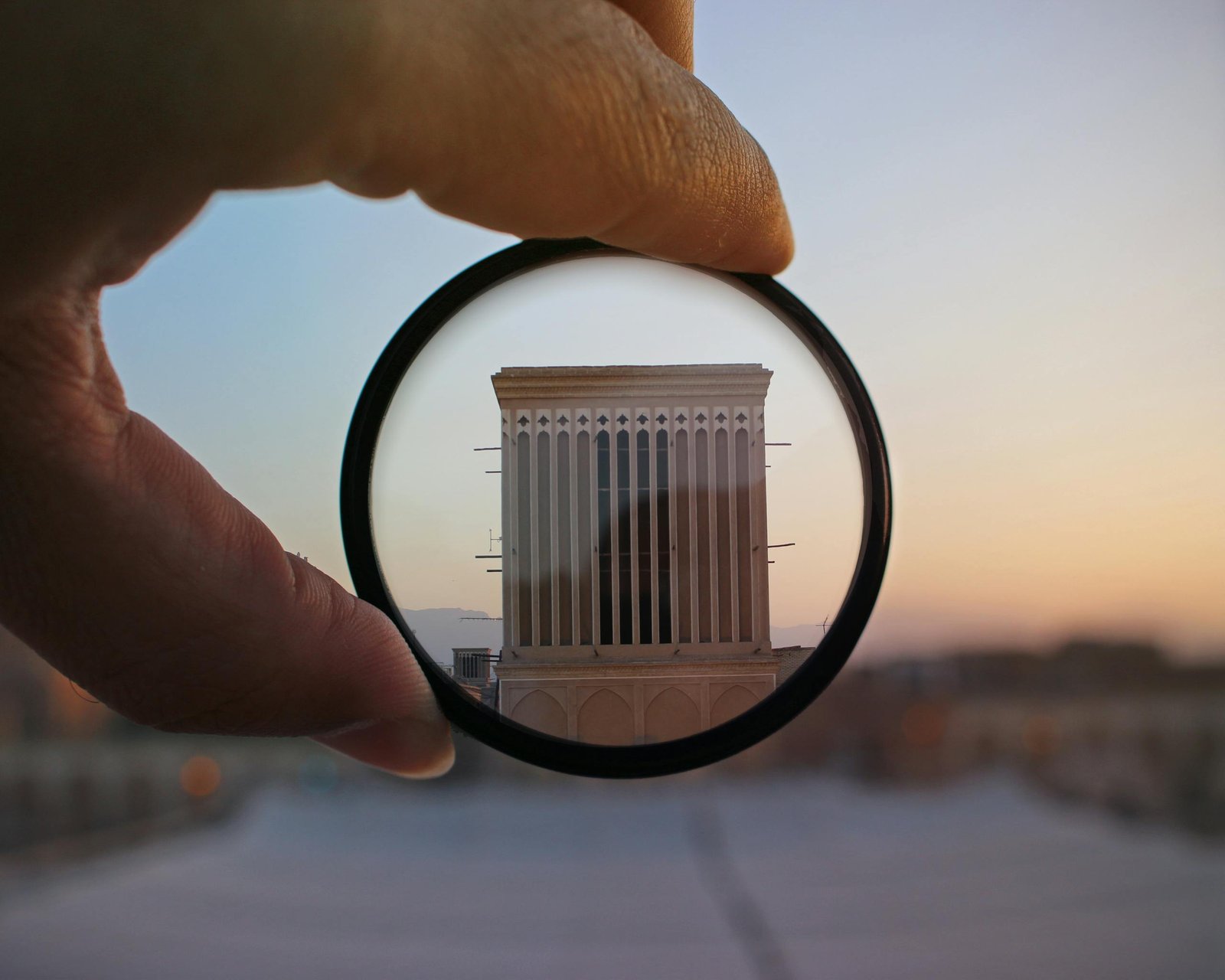 A hand holding a lens magnifies a building facade at sunset, highlighting architectural details in an abstract composition.